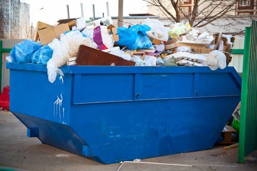 A large blue industrial rubbish skip filled with a mixture of waste materials including torn white and brown packing paper, broken cardboard boxes, plastic bags, and discarded packaging materials, situated outdoors on a paved area near a green metal fence. The skip is overflowing and stacked with waste, with some items spilling over the top edge, and is positioned in an area that appears to be part of a garden or residential outdoor space, possibly in Sanderstead. In the background, there are blurred trees and buildings, indicating an urban or suburban environment. The scene is lit by natural daylight, highlighting the various textures and colors of the waste materials and the metal surface of the skip, consistent with garden clearance or rubbish removal activities conducted by companies such as Garden Clearance Sanderstead.