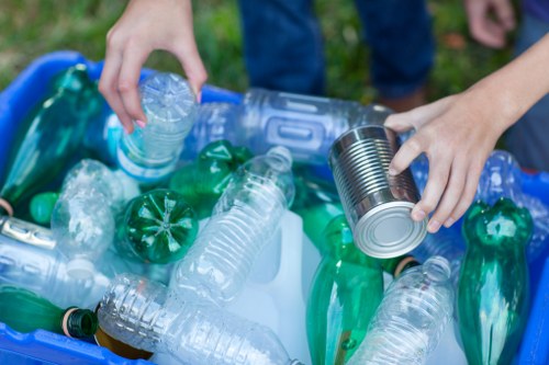 Close-up of a person's hands sorting recyclable waste, including clear plastic bottles, green plastic bottles, and a tin can, over a blue plastic bin filled with various plastic packaging and bottles. The background shows a grassy outdoor area, suggesting a garden or park setting. The person is wearing casual clothing, with a focus on the task of waste separation, supporting environmentally responsible rubbish disposal. This scene illustrates the importance of proper rubbish management and recycling, as offered by Garden Clearance Sanderstead, operating in the Sanderstead area, with a focus on rubbish removal services that handle garden and general waste collection to keep outdoor spaces tidy.