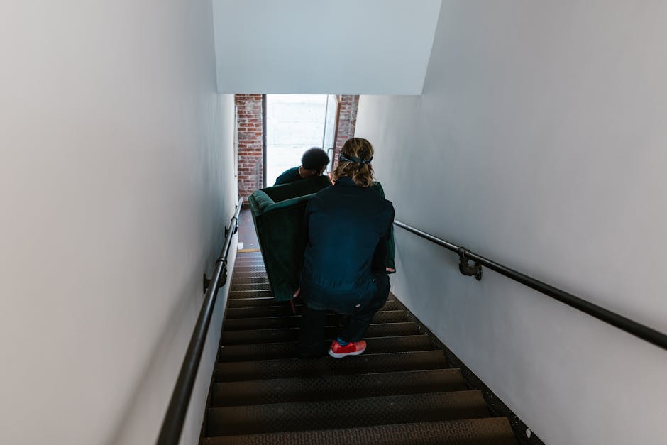Two individuals are descending a staircase in an indoor setting with plain white walls on either side and black handrails attached to the walls. The staircase has dark carpeting on the steps. One pers